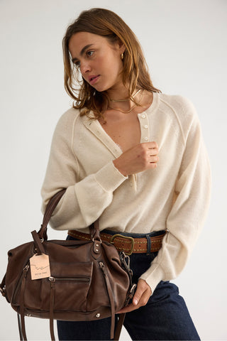 A woman wearing a cream ribbed knit henley cardigan, brown belt, and dark blue jeans, holding a brown leather shoulder bag against a plain light gray background.