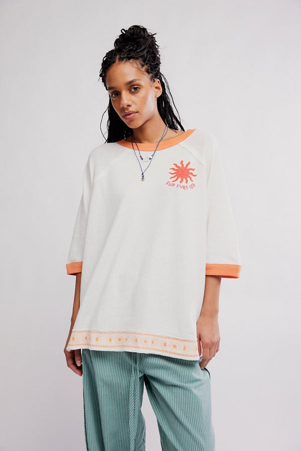Woman wearing a white oversized shirt with a red sun graphic and text, standing against a plain background.