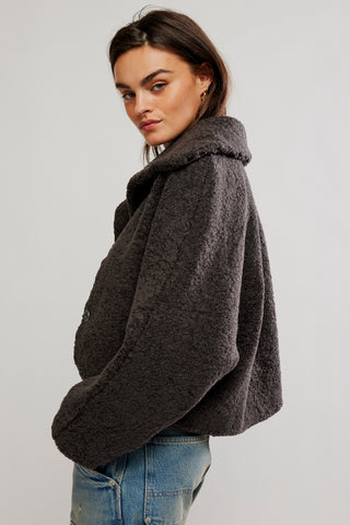 A woman wearing a dark brown textured teddy fleece jacket and blue jeans, photographed from a side-back angle against a plain light gray studio background.