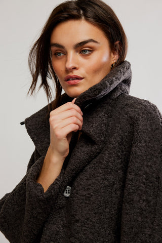 A close-up of a woman wearing a dark brown textured bouclé coat with a high fuzzy collar, adjusting the collar with her hand against a plain light gray background.