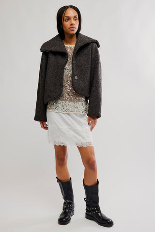 A model stands against a light gray studio background wearing a dark charcoal textured cropped jacket over a patterned top and white skirt, paired with black knee-high boots.