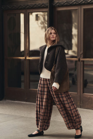 A woman stands on a sidewalk in front of large brown glass doors, wearing a dark brown shearling-style jacket over a cream ribbed sweater, loose brown and beige plaid wide-leg pants, and black flats.