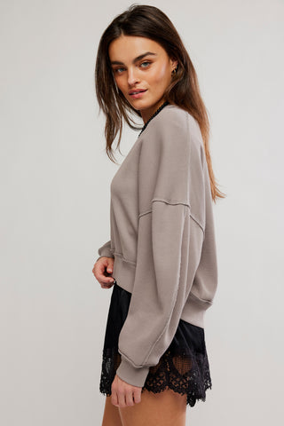 A woman modeling a taupe oversized crewneck sweatshirt with long sleeves and a relaxed fit, paired with black lace-trim shorts against a light gray studio background.