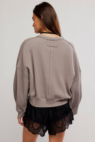 A woman shown from the back wearing a taupe-gray cropped sweatshirt and black lace-trimmed shorts against a light gray studio background.