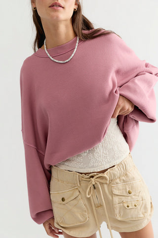 A woman wearing a dusty pink ribbed knit cropped sweatshirt, styled with a white textured top and beige drawstring shorts against a plain light background.