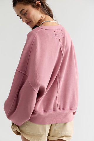 A woman wearing a dusty pink oversized crewneck sweatshirt with wide balloon sleeves, shown from a side-back angle against a light gray studio background.
