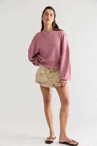 A woman modeling an oversized dusty rose crewneck sweatshirt, paired with beige cargo shorts and dark brown flip-flops, against a plain light gray studio background.
