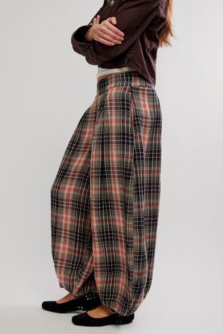 A person wearing loose, high-waisted plaid harem-style pants in black, red, and white, paired with a dark brown top and black flats, photographed against a plain light gray studio background.