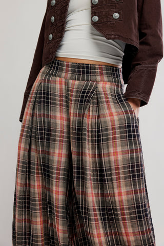 A close-up of a person wearing high-waisted, wide-leg plaid trousers in brown, red, and black, styled with a white ribbed top and a dark brown cropped jacket.