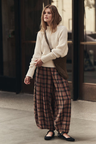 A woman stands on a city sidewalk wearing a cream ribbed turtleneck sweater, wide-leg brown and beige plaid pants, black flats, and a brown shoulder bag.