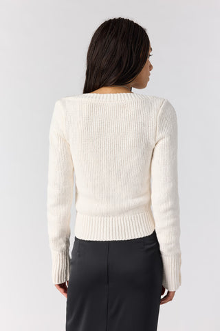 A woman shown from the back wearing a cream ribbed knit sweater and dark gray pants, photographed against a light gray studio background.