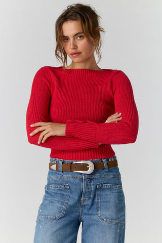 A woman wearing a bright red ribbed knit sweater with a wide boat neckline, paired with blue jeans and a brown belt, against a light gray studio background.