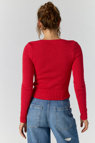 A woman viewed from the back wearing a bright red ribbed knit long-sleeve sweater and blue jeans against a light gray studio background.