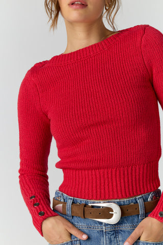 A close-up of a model wearing a bright red ribbed knit sweater with an off-the-shoulder neckline, paired with blue jeans and a brown belt.