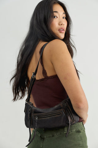 A woman wearing a dark brown ribbed camisole and olive green pants, shown from a side-back angle while wearing a black faux-leather crossbody bag with multiple zippered pockets.