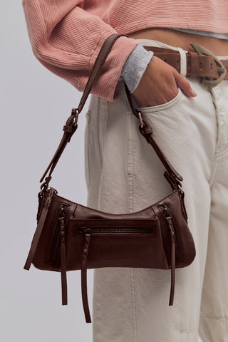A dark brown leather shoulder bag with multiple zippers and hanging tassels, shown being worn crossbody over the hip of a model wearing a pink ribbed sweater and cream pants.