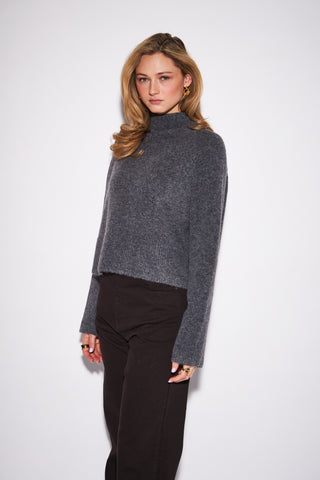 A woman modeling a dark gray cropped turtleneck sweater with long sleeves, paired with black trousers, against a plain light gray studio background.