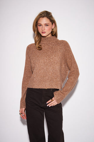 A woman modeling a cropped, long-sleeve knit turtleneck sweater in a heathered warm brown or camel color, paired with black pants, against a plain light gray studio background.