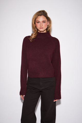 A woman modeling a dark burgundy ribbed knit mock neck sweater with long sleeves, paired with black pants, against a plain light gray studio background.