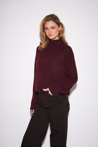 A woman modeling a dark burgundy knit mock neck sweater with black wide-leg pants, standing against a plain white studio background.