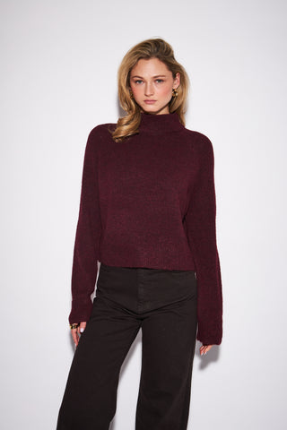 A woman modeling a cropped, long-sleeve ribbed knit turtleneck sweater in deep burgundy, paired with black wide-leg pants, against a plain light gray studio background.