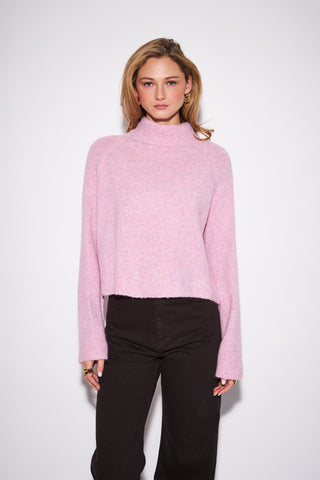 A woman modeling a light pink fuzzy knit mock-neck sweater with long sleeves, paired with black pants, against a plain light gray studio background.