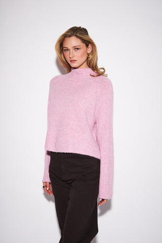 A woman modeling a light pink fuzzy mock-neck sweater paired with black trousers, photographed against a clean white studio background.