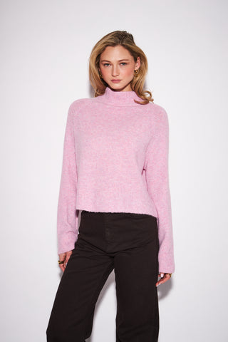 A woman modeling a light pink fuzzy mock-neck sweater paired with black pants, standing against a plain white studio background.