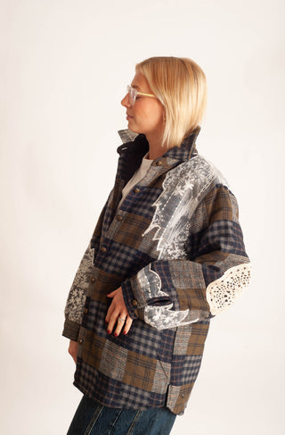 A side-profile view of a model wearing a patchwork plaid jacket with brown, navy, and gray tones, featuring distressed details and lace applique on the sleeve, set against a plain off-white background.