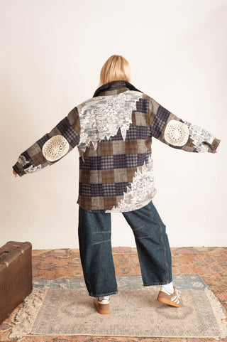 A person viewed from behind wearing a patchwork patchwork jacket with brown plaid, white lace, and circular crochet details, paired with wide-leg blue jeans and tan shoes, standing on a patterned rug.