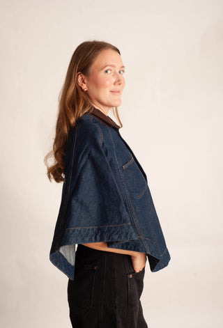 A woman wearing a dark blue cropped denim jacket with a brown collar, shown in profile against a plain off-white studio background.
