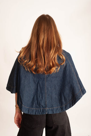 A woman viewed from behind wearing a dark blue denim cape-style top with wide short sleeves and black pants, against a plain off-white background.