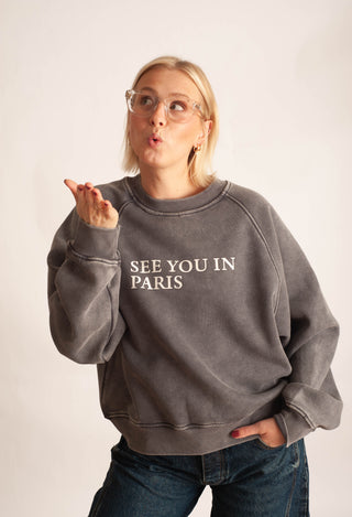 A woman wearing a washed charcoal gray sweatshirt with 'SEE YOU IN PARIS' printed in white on the chest, paired with blue jeans, posing against a light off-white background.