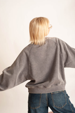 A person shown from behind wearing a washed charcoal gray oversized sweatshirt and blue jeans against a plain off-white background.