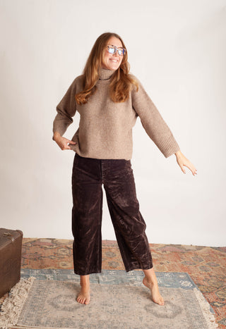 A woman modeling a beige knit mock-neck sweater and dark brown wide-leg velvet pants, standing barefoot on a patterned rug against a plain white wall.