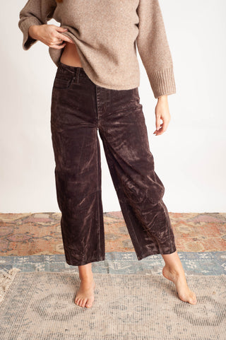 A person wearing dark brown corduroy cropped pants and a beige knit sweater, standing barefoot on a textured rug against a plain light wall.