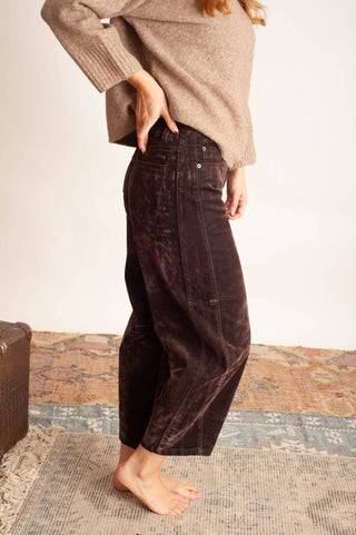 A person wearing dark brown wide-leg corduroy pants and a beige knit sweater, standing barefoot on a patterned rug against a plain light wall.
