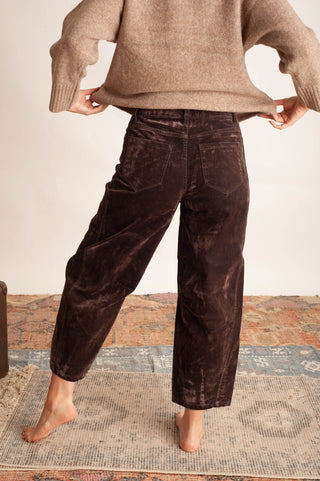 A person shown from the back wearing dark brown velvet wide-leg cropped pants and a beige knit sweater, standing barefoot on a patterned rug against a plain light wall.