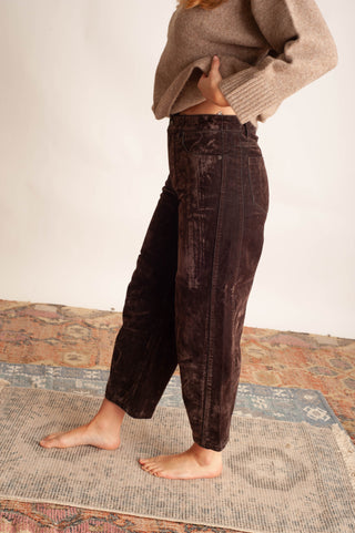 A person wearing dark brown corduroy wide-leg pants and a beige knit sweater, standing barefoot on a patterned rug against a plain off-white wall.