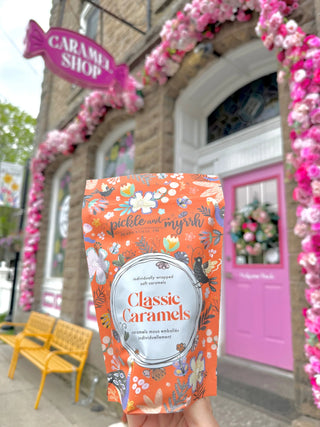 A hand holds a bag of 'Classic Caramels' by the brand 'pickle and mysh' in front of a whimsical shop facade decorated with pink flowers and a bright pink door.