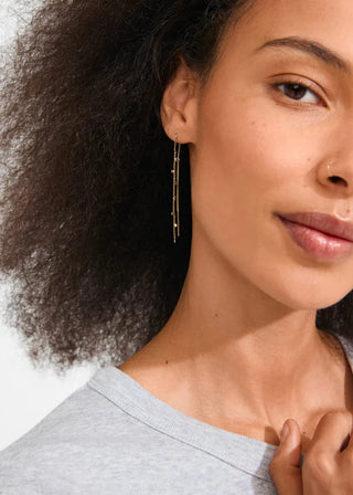 A close-up of a woman wearing a delicate gold dangle earring with multiple thin vertical bars, paired with a light gray crewneck top.