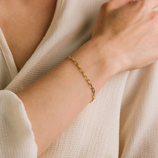 A person wearing a gold paperclip chain bracelet on their wrist.