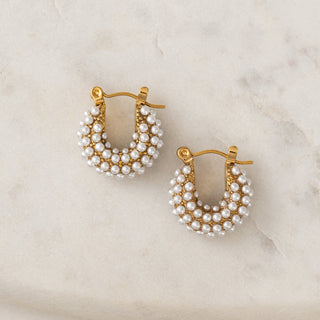 A pair of gold-colored hoop earrings with white dotted pearls, displayed on a textured background.
