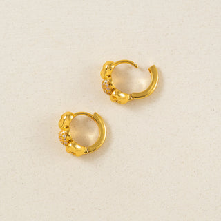 A pair of gold huggie hoop earrings with a textured, organic raised design and small clear crystal accents, displayed on a light beige textured background.