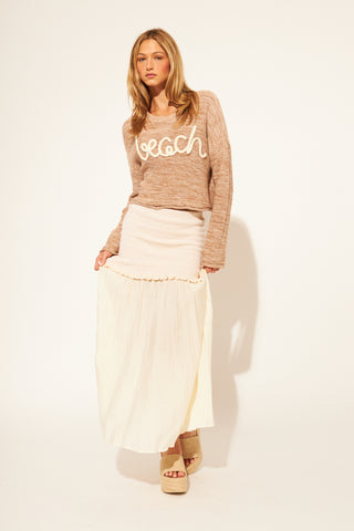 Woman wearing a brown 'beach' sweater and white skirt on a plain background