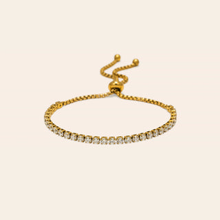 A gold adjustable bracelet featuring a single row of clear pavé-set round stones, displayed on a solid light beige background.