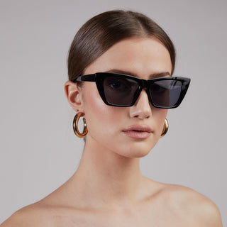 Woman wearing black sunglasses and gold hoop earrings against a gray background