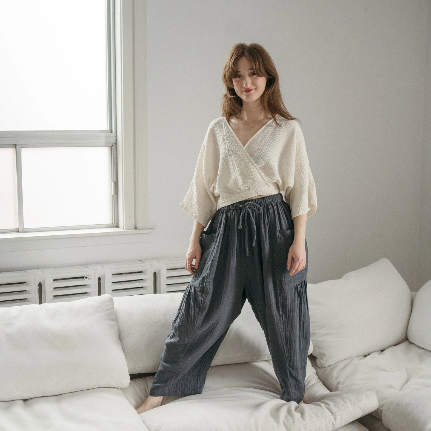 Woman wearing a white blouse and dark pants standing in a room with a white couch and window.