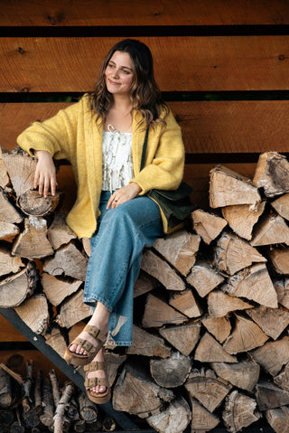women wearing a yellow cardigan sitting on a bench of logs in front of a barn. 