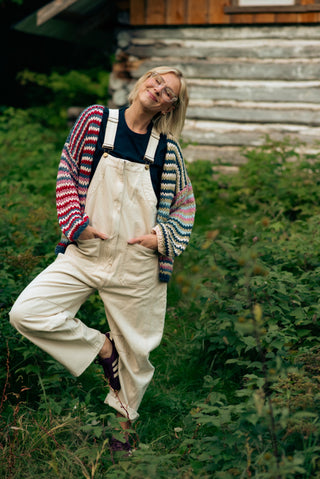 women wearing overalls and a rainbow cardigan in a field of grass in front of a sugar shack. 